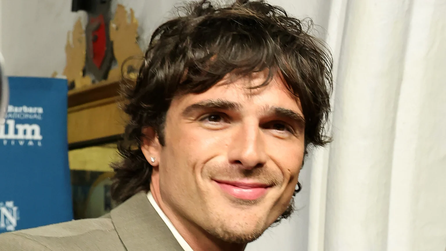 Close-up of actor Jacob Elordi smiling slightly, showing his dark wavy hair, stubble, and stud earrings, with a partial blue 'film' sign visible in the blurred background.