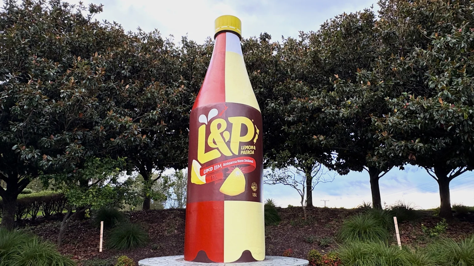 A giant L&P soda bottle, half reddish-brown and half yellow, stands outdoors surrounded by green trees under a cloudy sky.