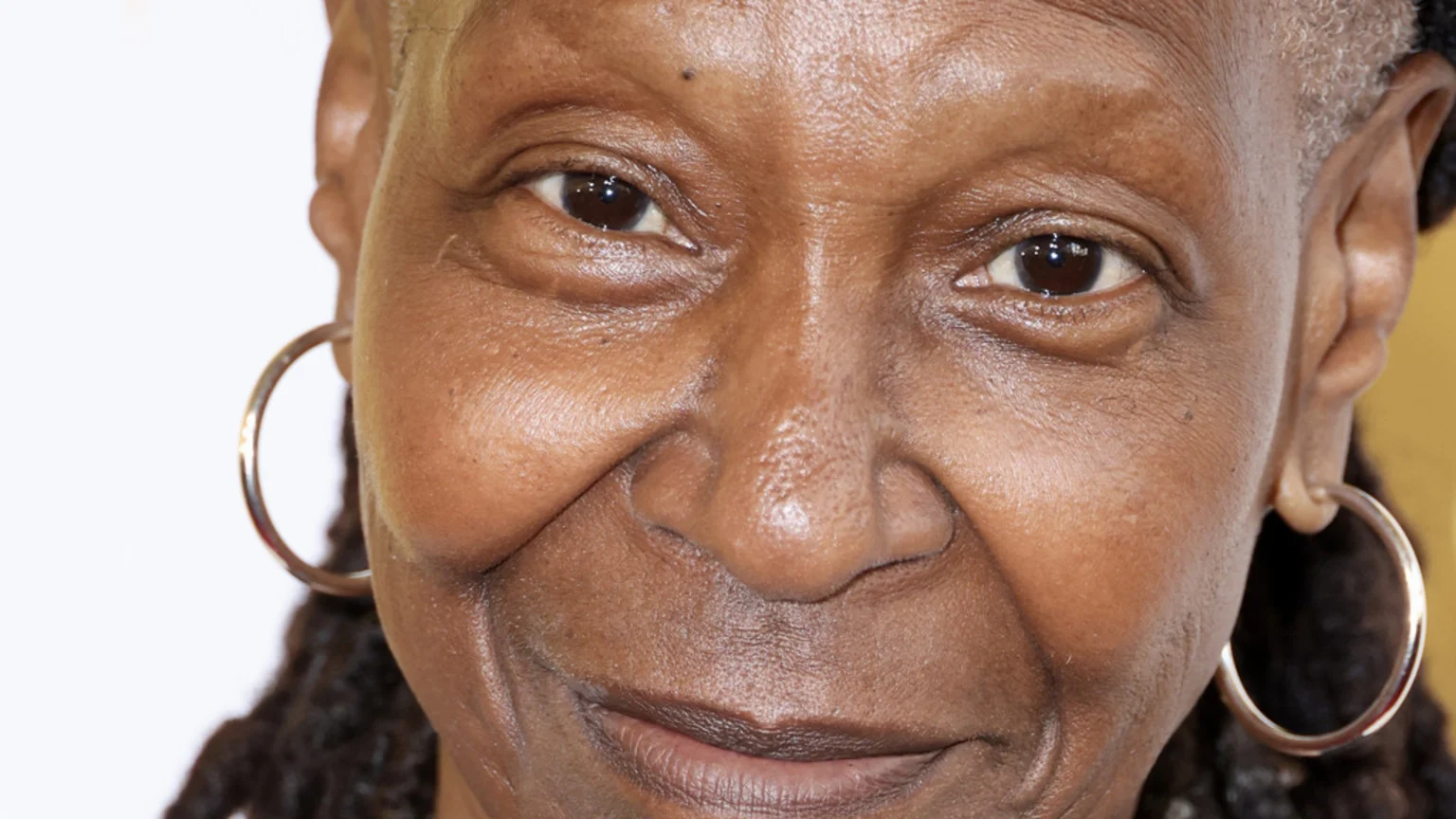 Close-up portrait of Whoopi Goldberg smiling faintly, showing wrinkles around her dark eyes and cheeks, wearing hoop earrings.