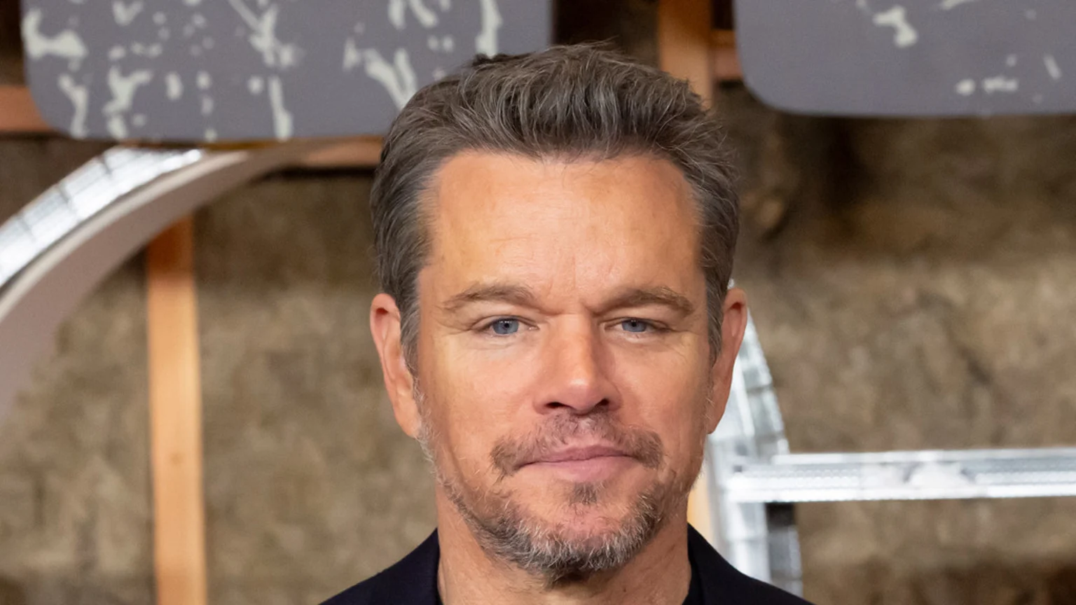 Close-up of actor Matt Damon with salt-and-pepper hair, blue eyes, and a light beard, looking directly at the camera.