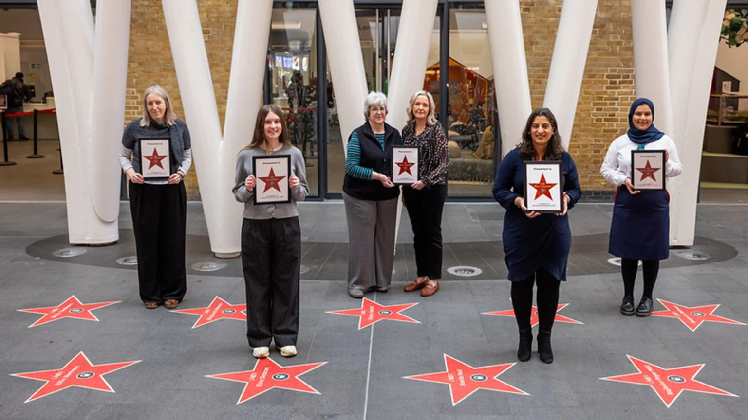 Six women stand in an indoor space, each holding a framed award with a red star, with star decals on the floor around them.