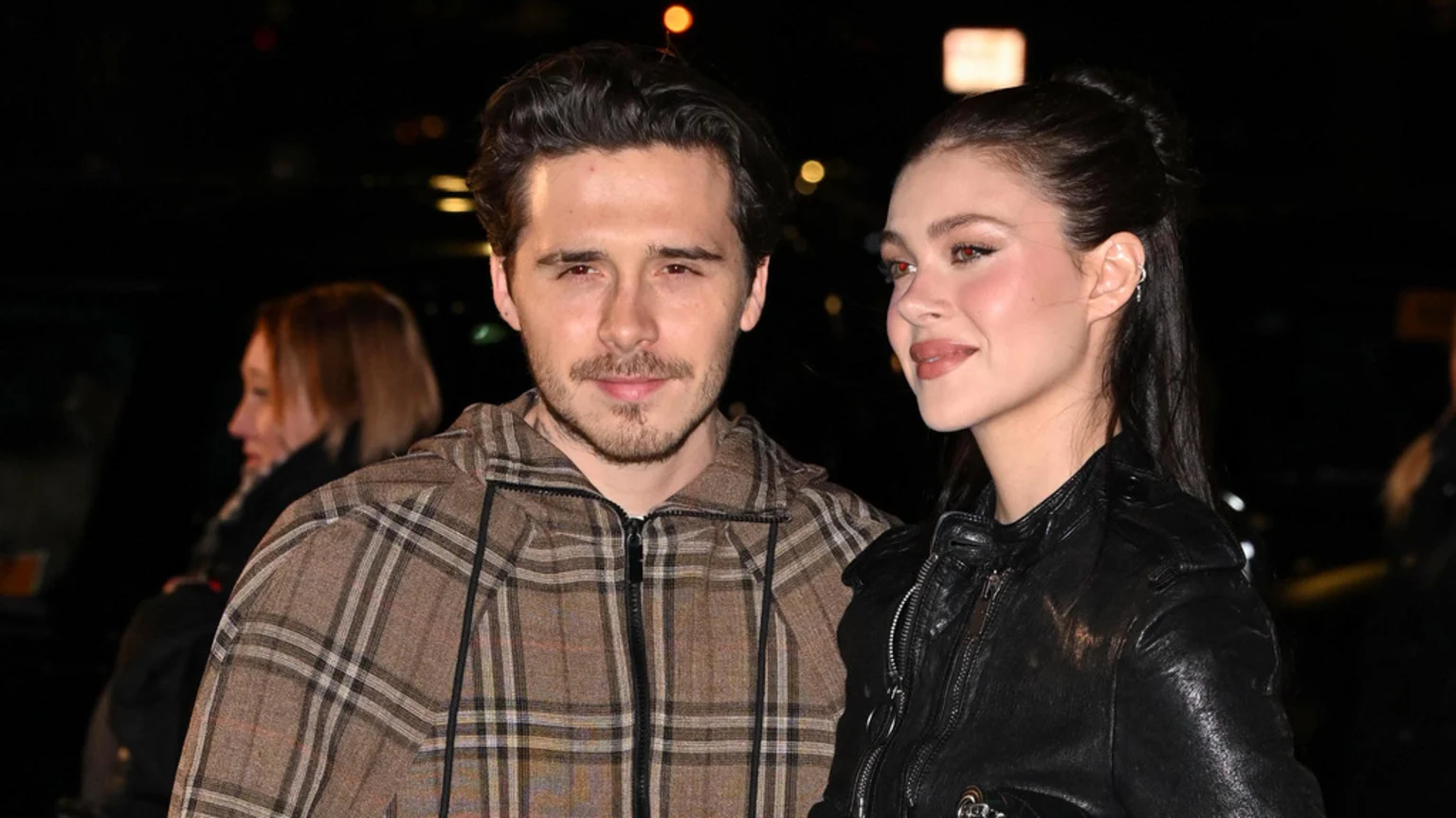 A candid nighttime portrait of Brooklyn Beckham and Nicola Peltz outdoors on an urban street or event entrance. Brooklyn wears a brown plaid hooded jacket, facing camera; Nicola in a black leather jacket looks to her right. An adult person appears blurred in background.