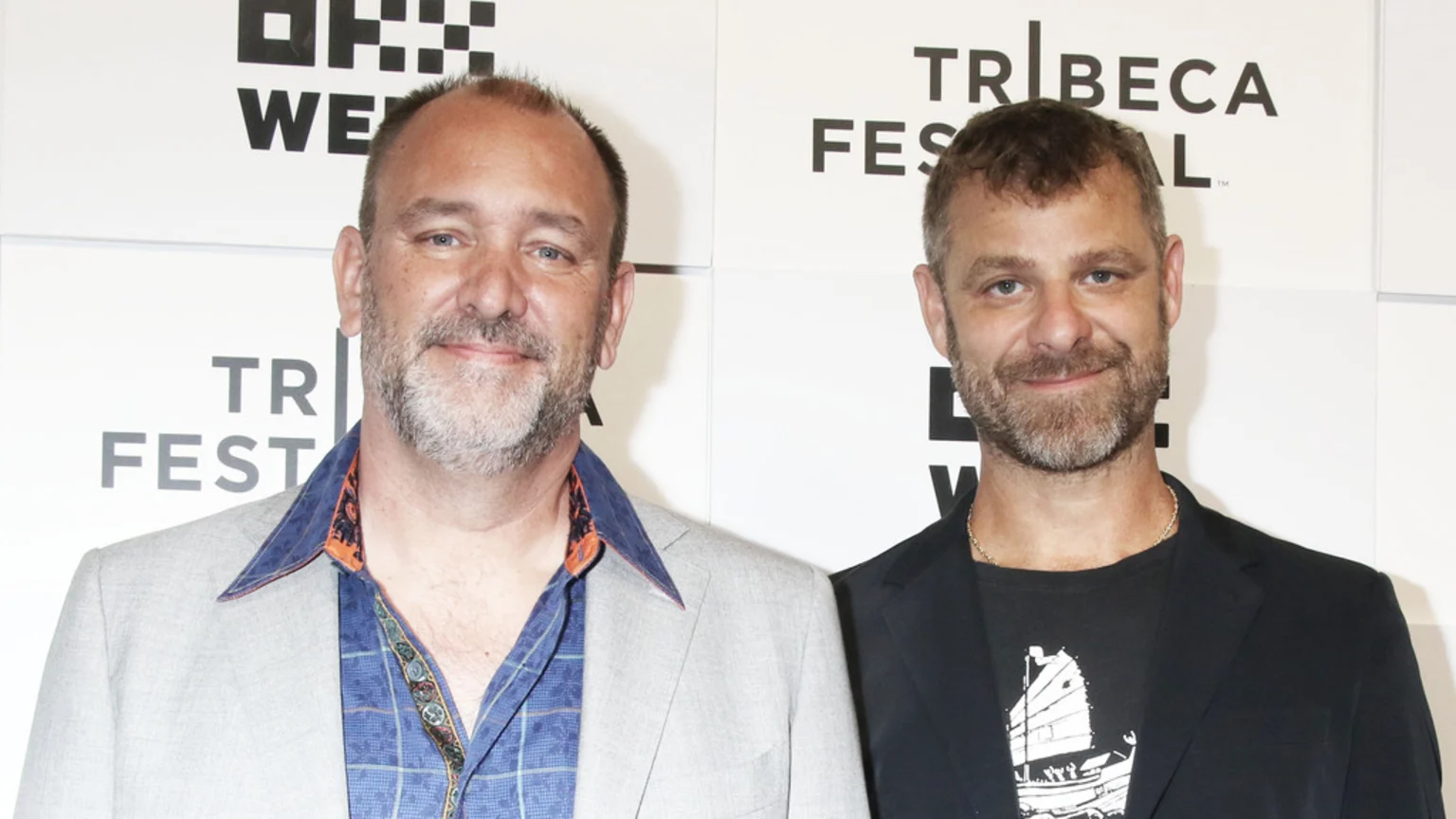 Two smiling men with beards stand against a white backdrop with black text. The man on the left wears a light blazer over a patterned shirt, while the man on the right wears a dark blazer over a graphic tee and a gold chain.