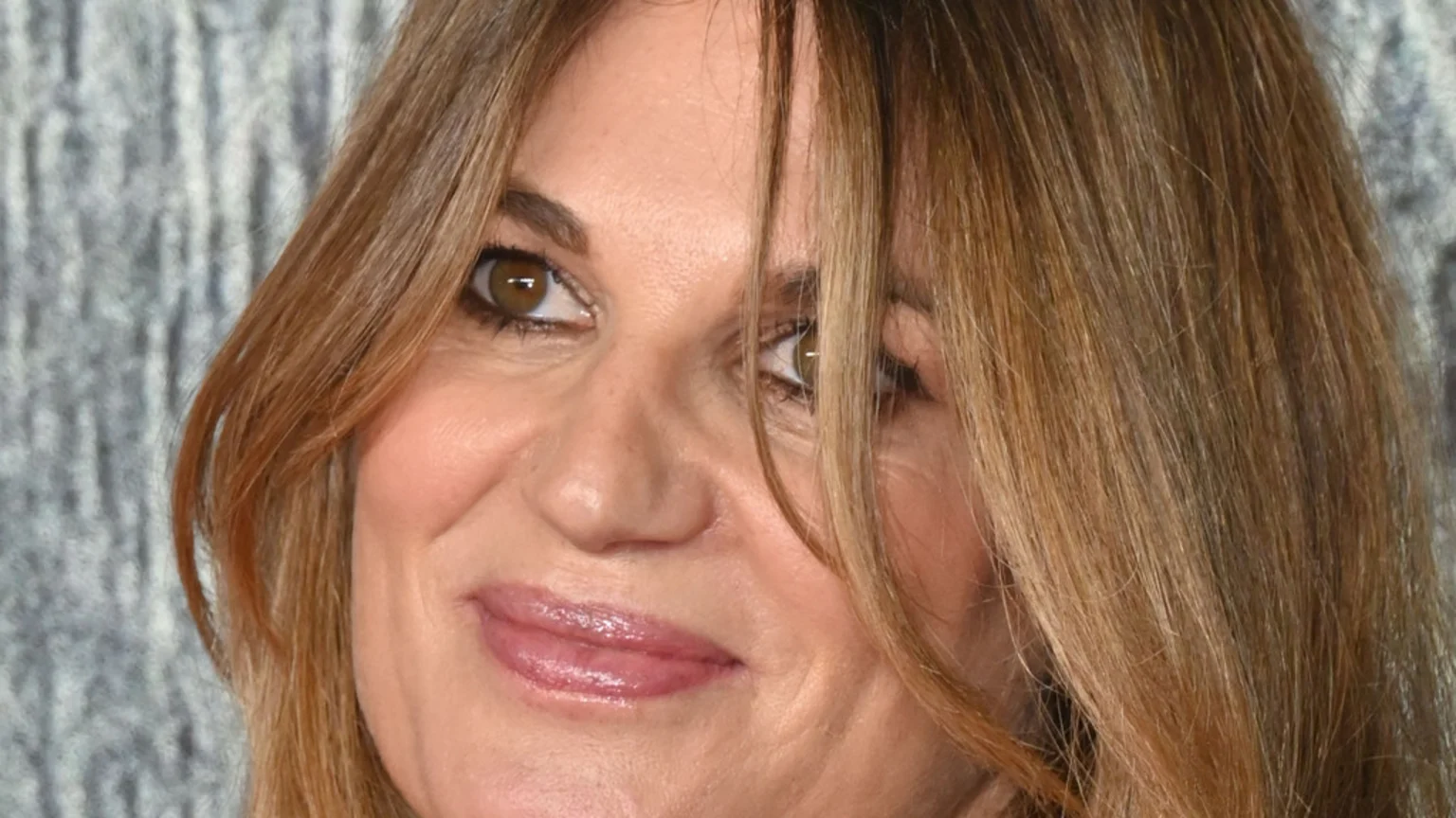 Close-up of a woman with light brown hair, brown eyes, and glossy pink lips, smiling slightly against a blurry grey background.
