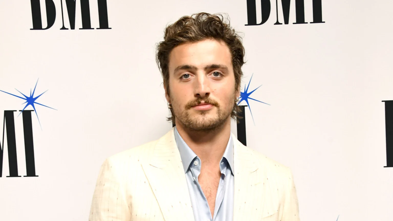 A young man with curly brown hair, a mustache, and a beard, wearing a light-colored suit jacket and a light blue shirt, looks at the camera against a white "BMI" logo background.