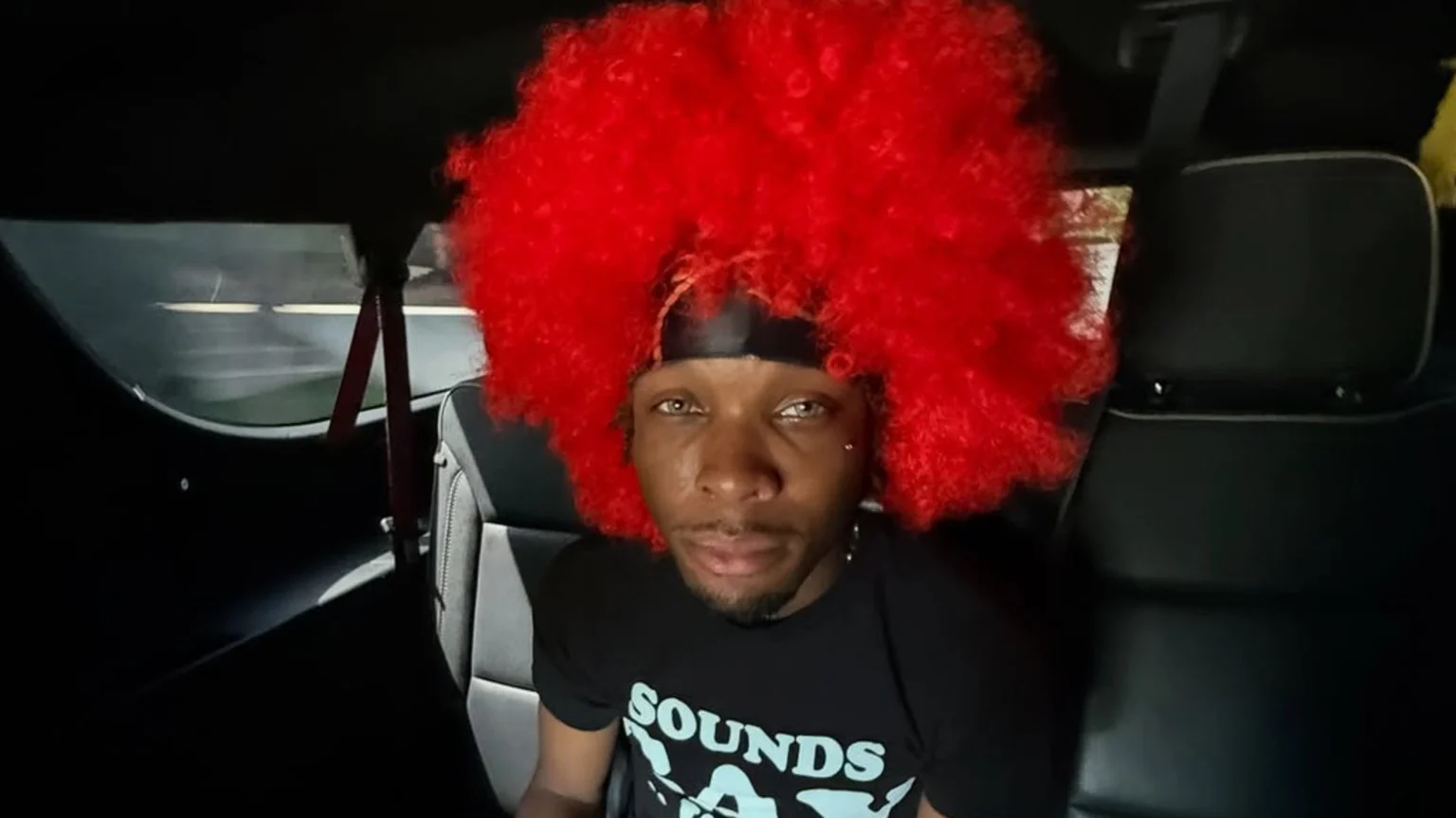 A person with dark skin, light eyes, and a large red afro wig looks directly at the camera while sitting in a dark car interior.