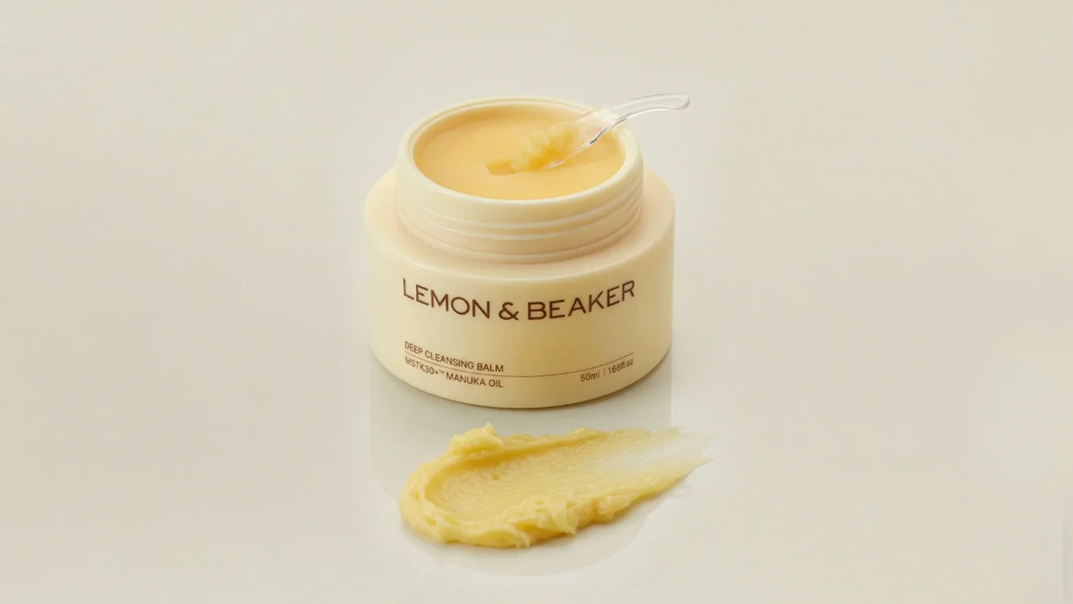 An open jar of Lemon & Beaker Deep Cleansing Balm with Manuka Oil, a spatula in the light yellow balm, and a swatch of the balm next to it on a light background.