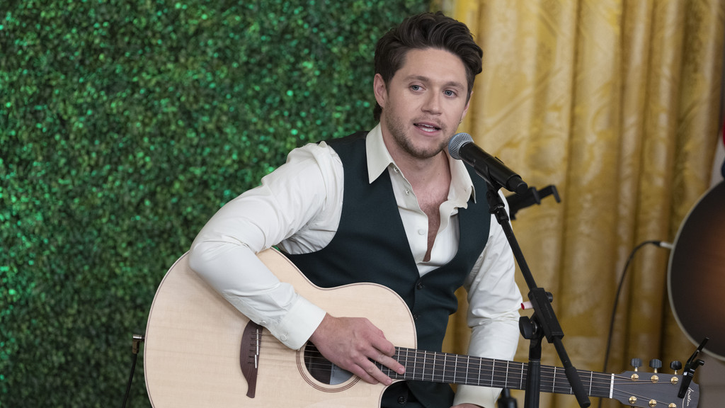 Niall Horan, wearing a white shirt and dark vest, sings into a microphone while playing an acoustic guitar, with a green wall and golden curtain in the background.