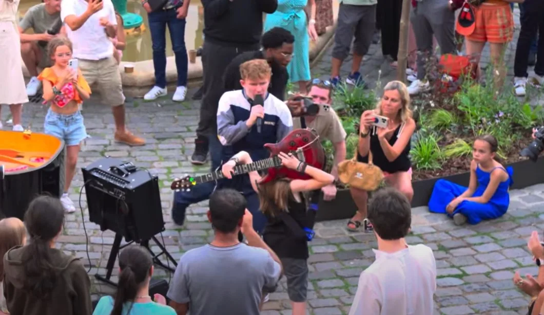 11 year old boy with long brunette hair plays red electric guitar behind his head on a busy street in Paris.