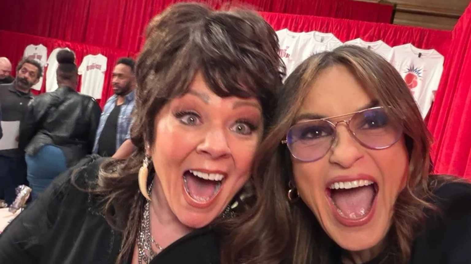 Melissa McCarthy and Mariska Hargitay take an excited selfie with wide open mouths, with a red curtain and white t-shirts in the background.