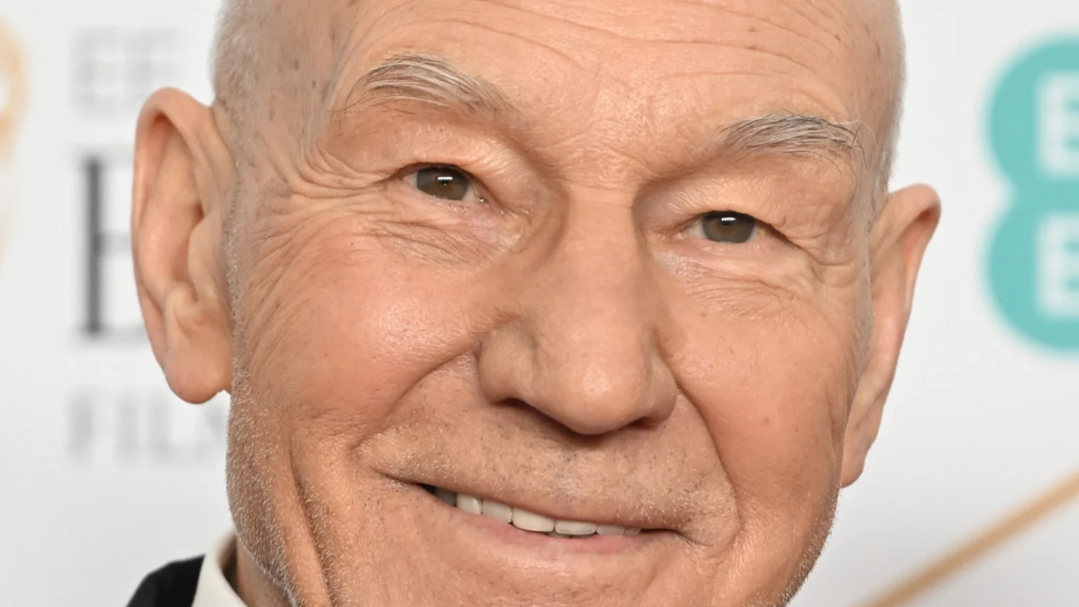 Close-up of an older, bald man with a light five o'clock shadow smiling, with brown eyes and grey eyebrows, against a blurry white background.