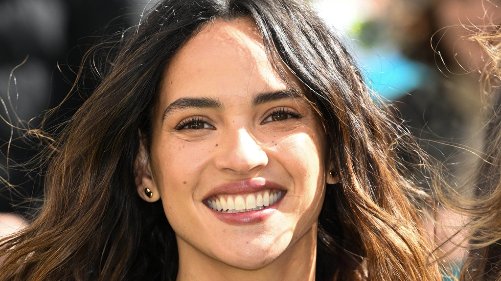 A close-up shows a smiling woman with dark brown hair and freckles, wearing gold stud earrings, her hair flowing around her face as she looks forward with a bright expression.