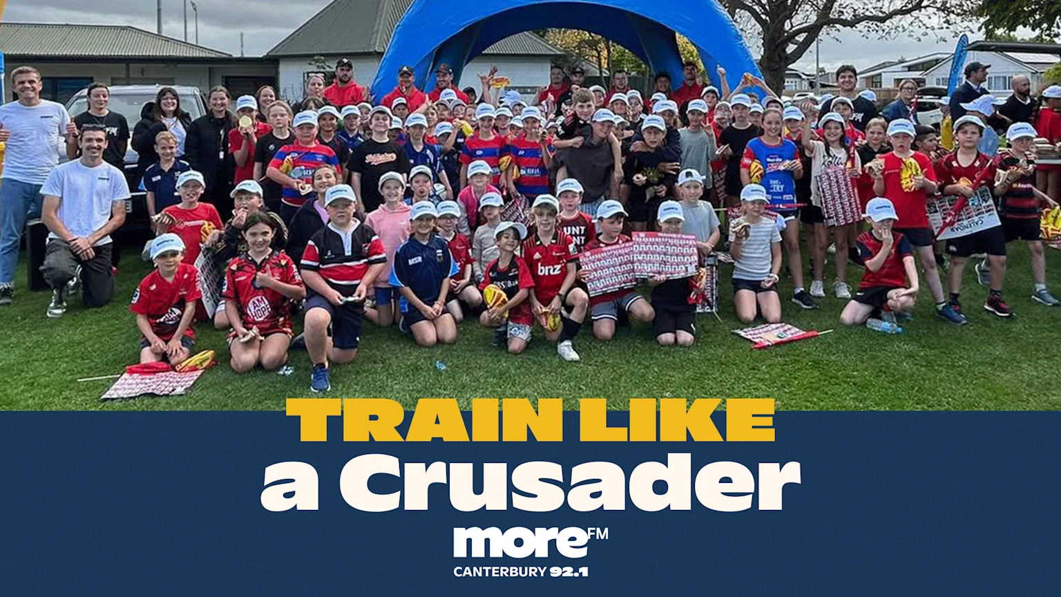 A large group predominantly of children wearing light blue baseball caps and sports jerseys poses on a grassy sports field. A blue inflatable arch and promotional banners with sponsor logos like Canterbury and BNZ appear in the background under an overcast sky.