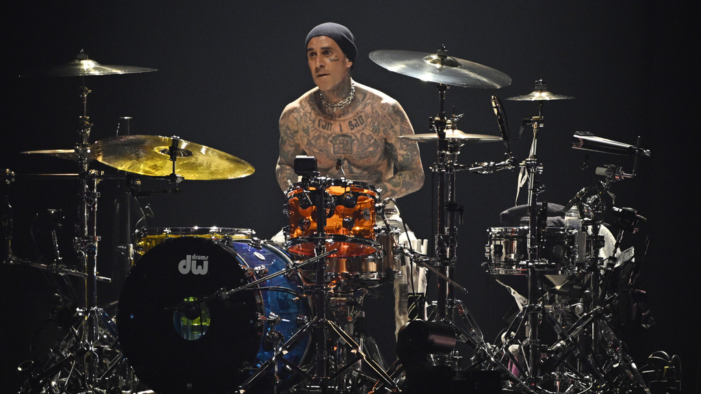 Travis Barker, shirtless and heavily tattooed, plays a multi-colored drum set on a dark stage.