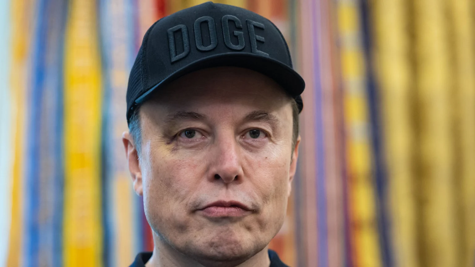 Close-up of Elon Musk wearing a black "DOGE" baseball cap, looking forward, with a blurred colorful background.
