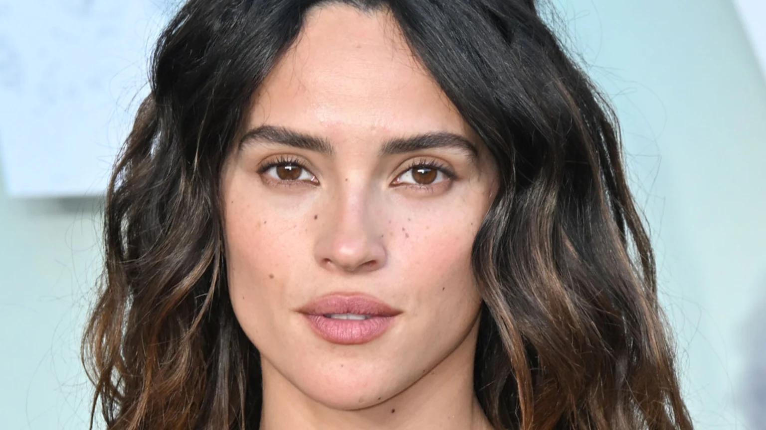 A close-up of actress Adria Arjona, featuring her dark wavy hair, brown eyes, freckled skin, and lips with light pink lipstick, as she looks directly at the camera.