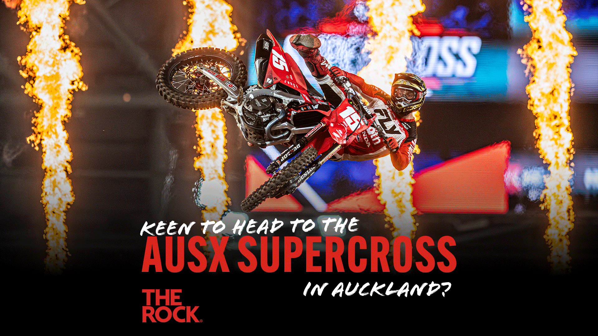 A motocross rider in red gear performs an aerial stunt on a red dirt bike between two columns of flames, with overlay text promoting 'AUSX SUPERCROSS IN AUCKLAND?'.