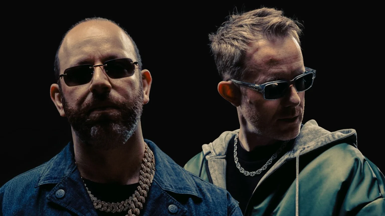 Two men in sunglasses and jackets pose against a dark background; one bearded with a gold chain and denim jacket, the other with short hair, a silver chain, and a green hoodie jacket.