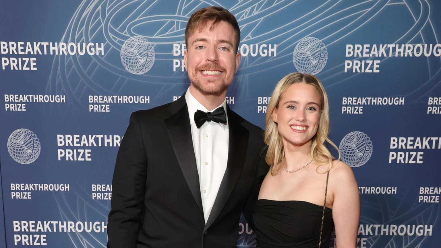 A smiling man in a black tuxedo and a blonde woman in a black strapless dress pose together in front of a blue backdrop featuring the "Breakthrough Prize" logo.