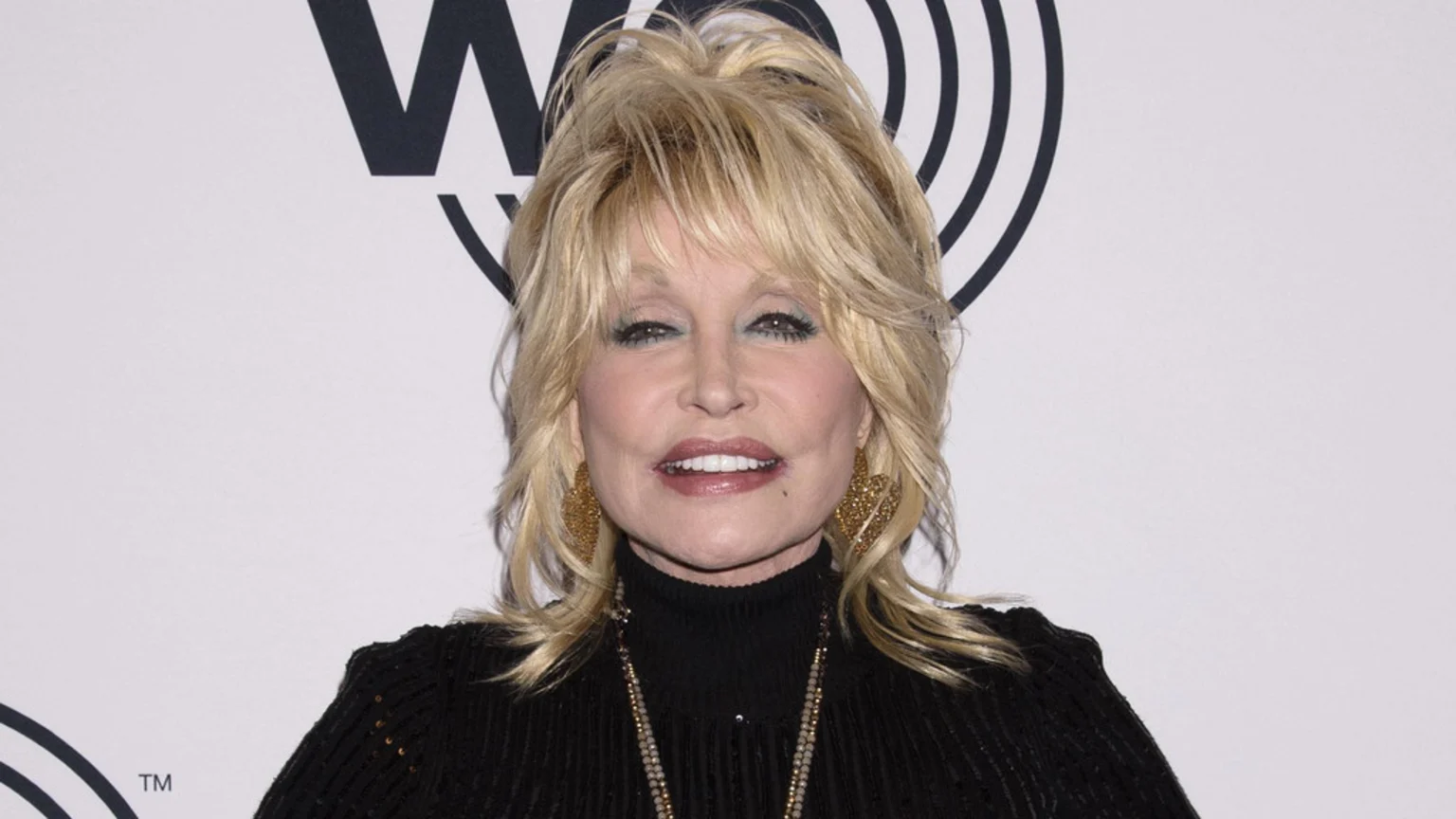 Dolly Parton smiles at the camera, wearing a black turtleneck, gold earrings, a beaded necklace, and heavy makeup with teal eyeshadow and pink lipstick, against a white background with a partial black logo.