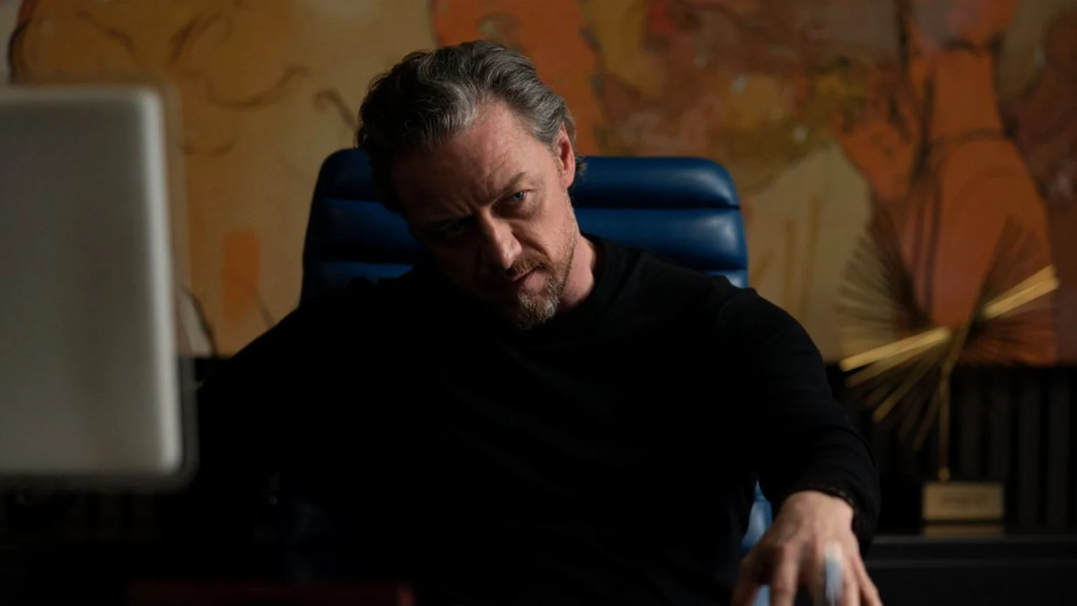 James McAvoy, with graying hair and beard, sits in a blue chair wearing a black shirt, looking intensely at the camera with his head slightly tilted.