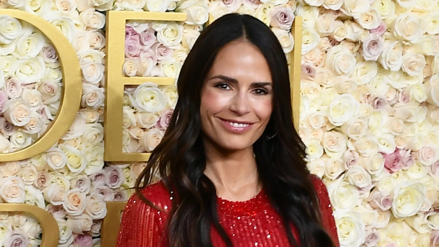 Jordana Brewster, with long dark hair and a red sequined top, smiles faintly in front of a wall of light-colored roses and gold letters spelling out parts of "GOLD".