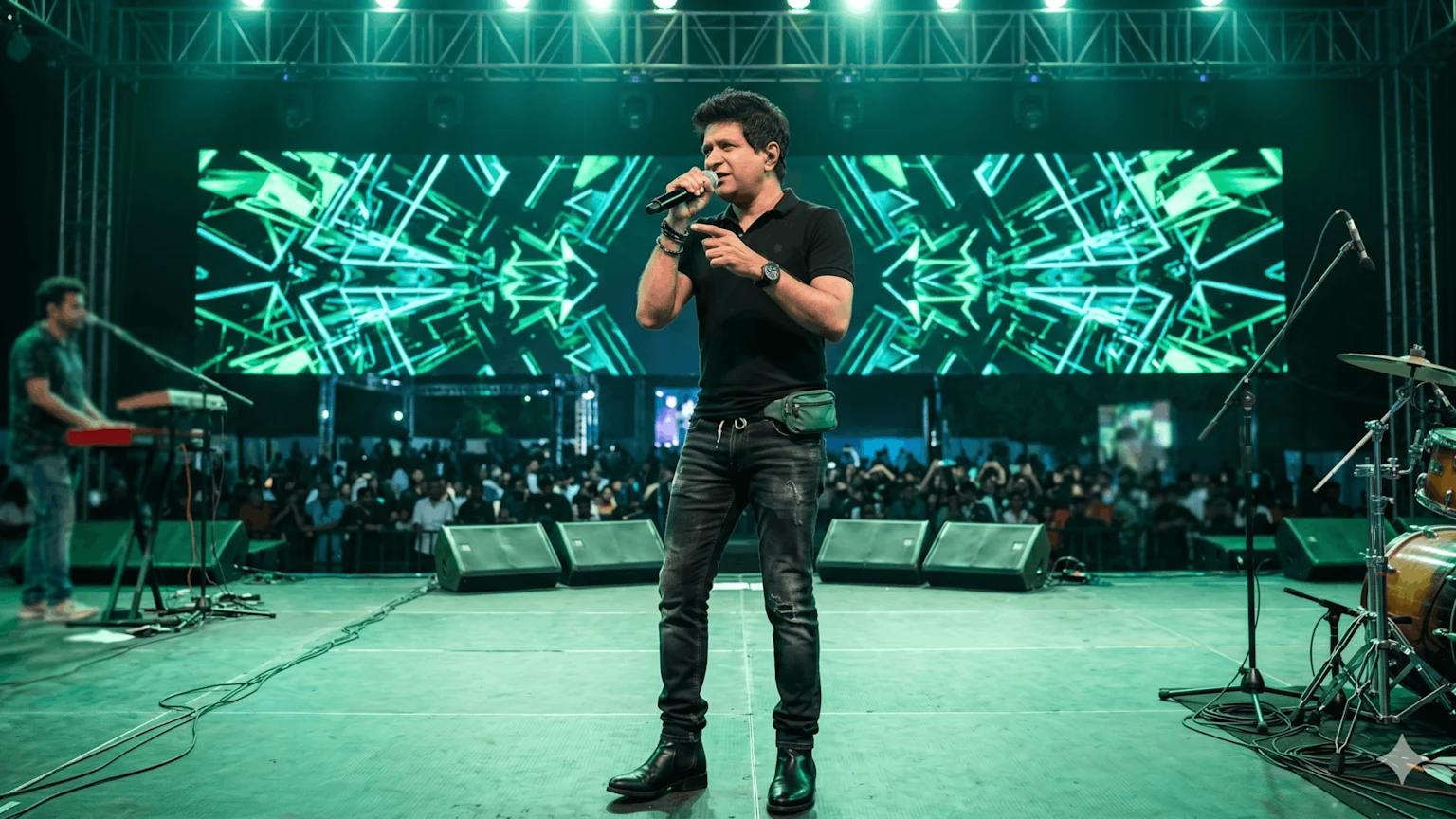 A male singer performs on stage with a microphone, a vibrant green abstract LED screen behind him, a keyboardist, drum kit, and an audience in the background.