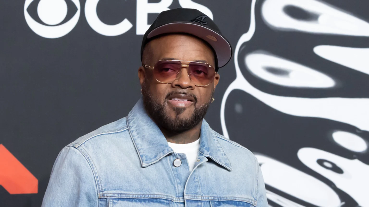 A man with a beard, wearing pink-tinted sunglasses, a black cap, and a denim jacket, smiles slightly in front of a dark grey background with white logos, including CBS.