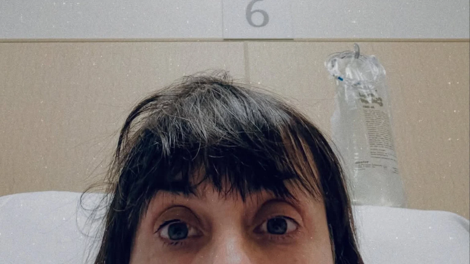 A close-up shows a person with messy dark hair and wide blue eyes, lying in a bed with an IV bag and the number 6 visible in the background.