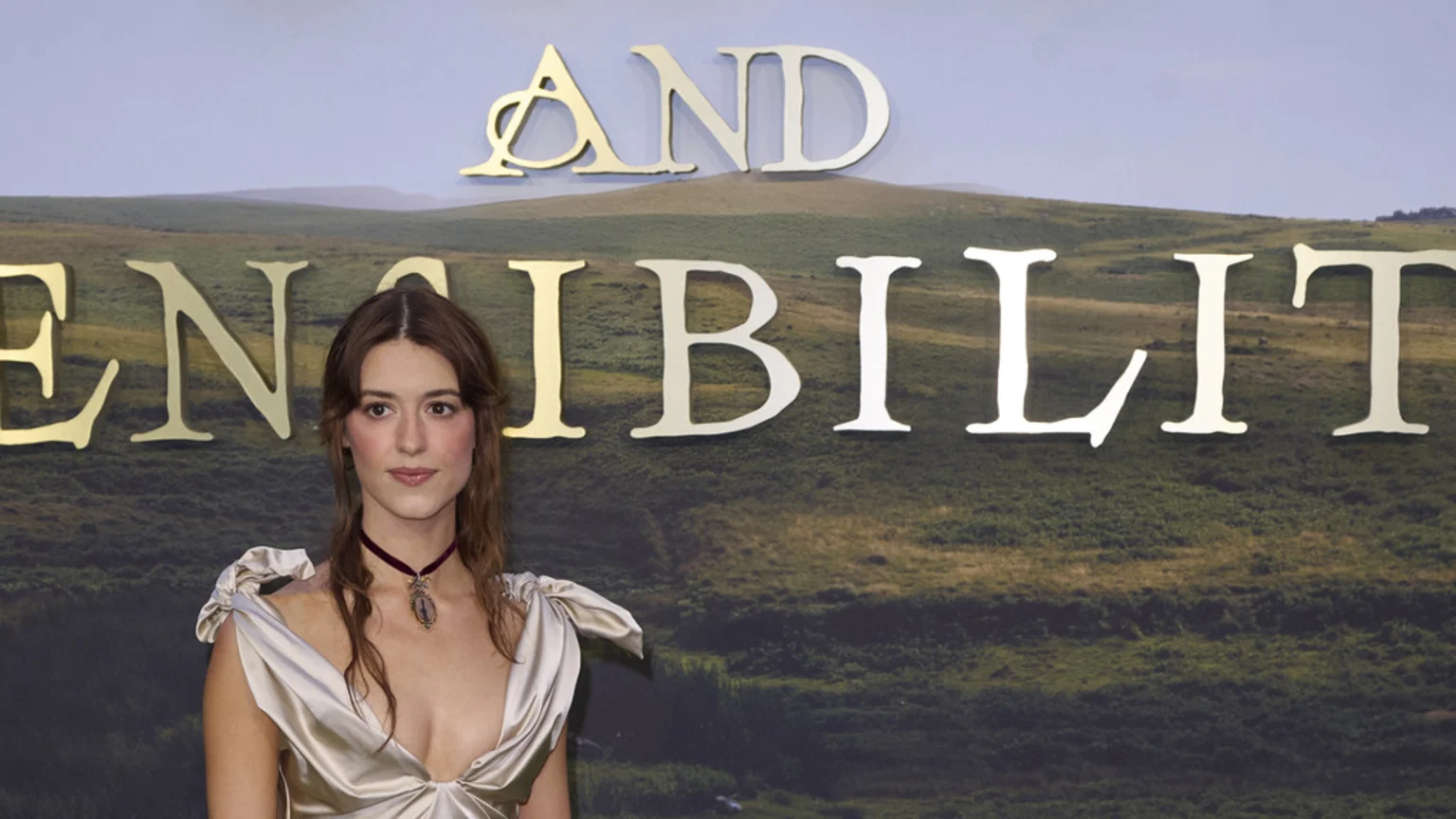 Actress Daisy Edgar-Jones, in a low-cut dress and choker, stands before a backdrop of green hills featuring the golden title 'SENSE AND SENSIBILITY'.