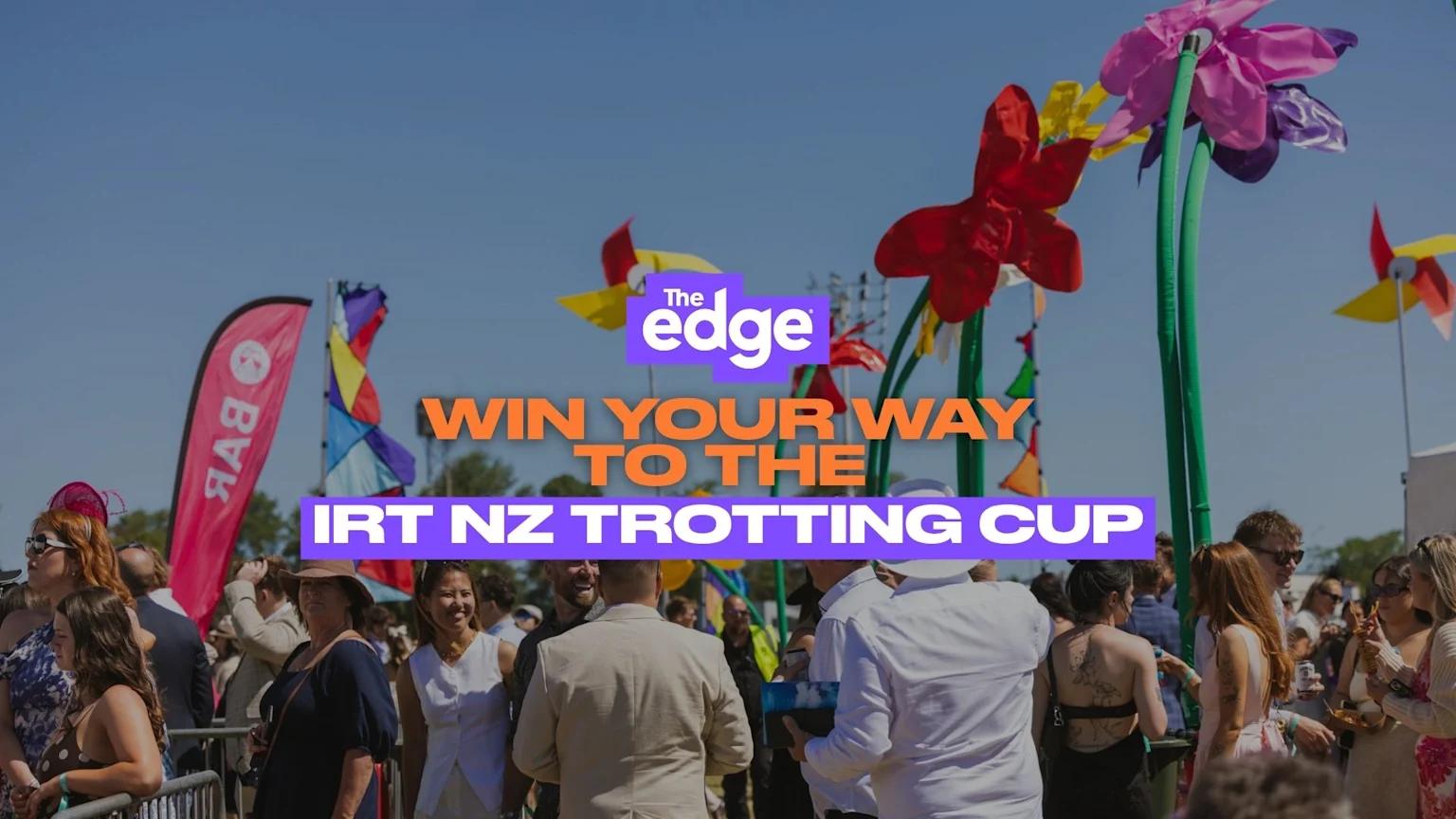 A sunny outdoor event scene with a crowd of people, colorful flags, and large floral decorations, featuring the text "The Edge WIN YOUR WAY TO THE IRT NZ TROTTING CUP."