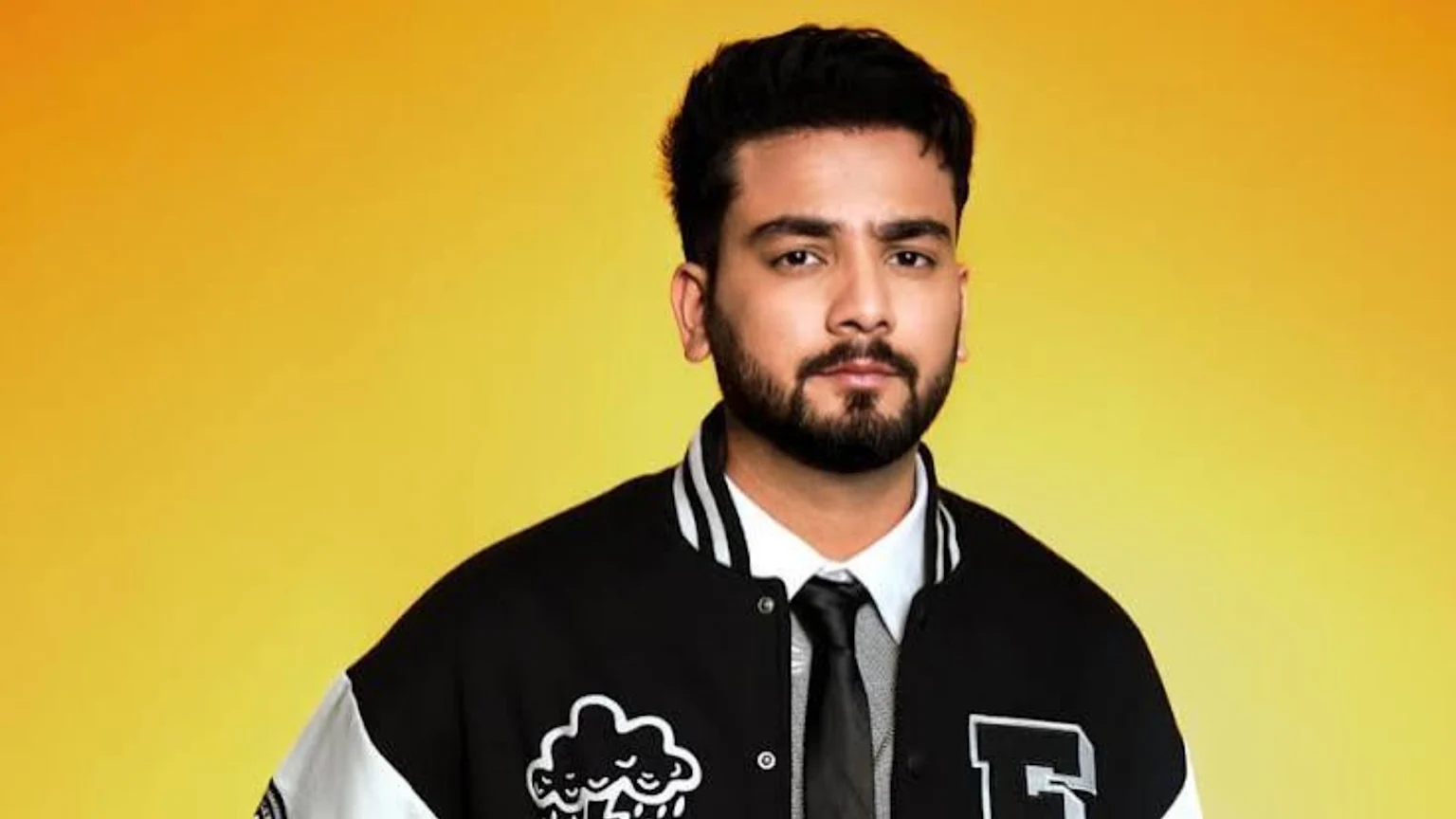 A man with a beard and dark hair wears a black and white varsity jacket featuring a cloud graphic and the letter 'E', over a white shirt and dark tie, looking at the camera against a yellow and orange gradient background.