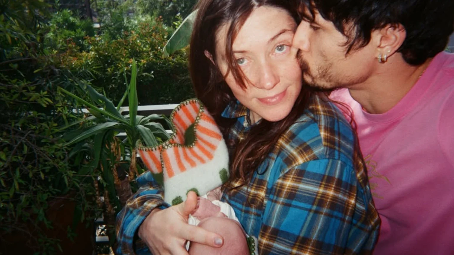 A man with a beard kisses a smiling woman's cheek as she holds a baby wrapped in an orange and white striped knitted item, with lush green plants in the background.