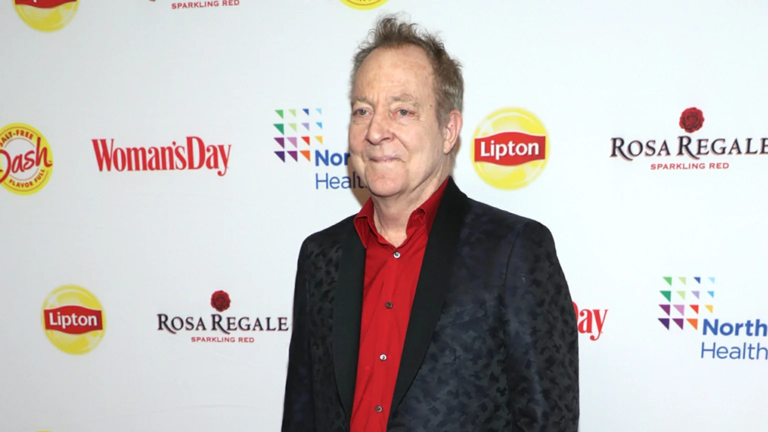 A smiling man in a red shirt and dark patterned blazer stands against a white backdrop displaying sponsor logos including Lipton and Woman's Day.