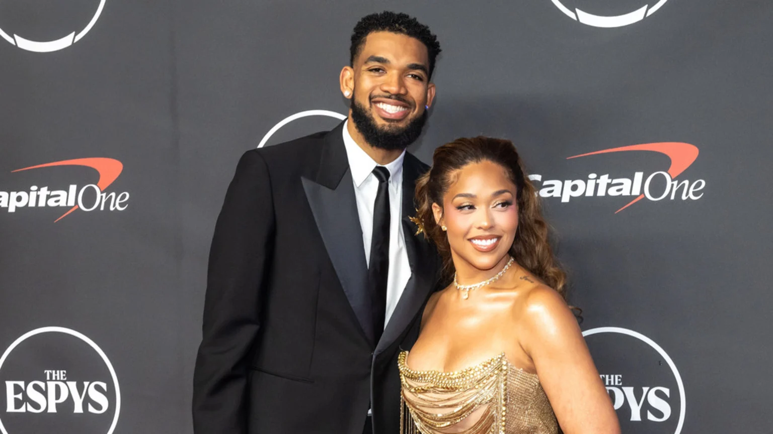 A smiling man in a black tuxedo stands with a smiling woman in a gold strapless dress against a black backdrop with Capital One and ESPYS logos.