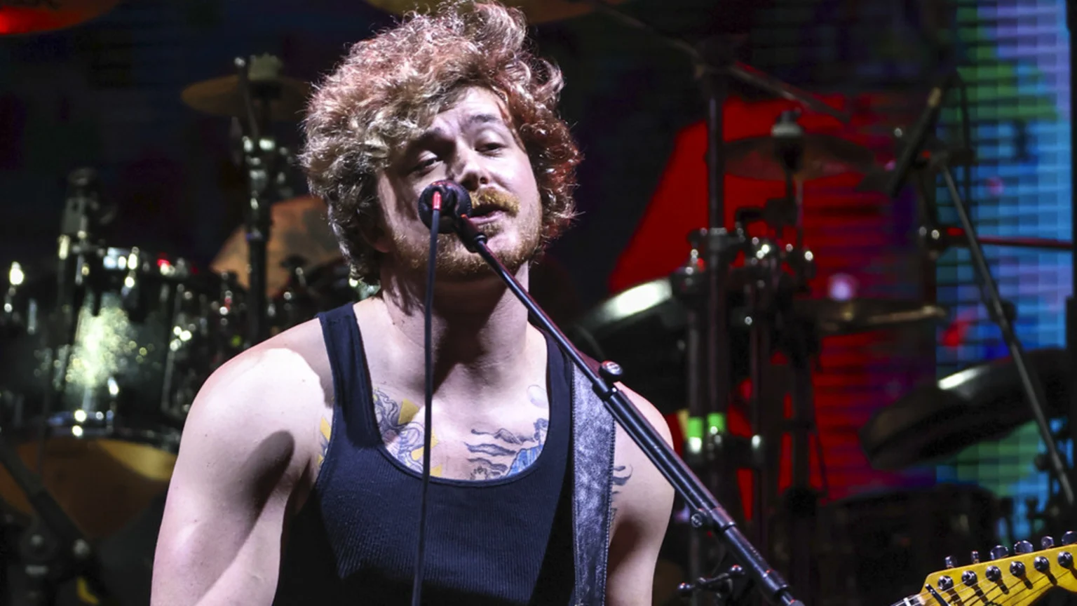 A male musician with curly brown hair and a beard sings into a microphone on stage, wearing a black tank top and showing tattoos, with blurred drum kits and colored stage lights behind him.