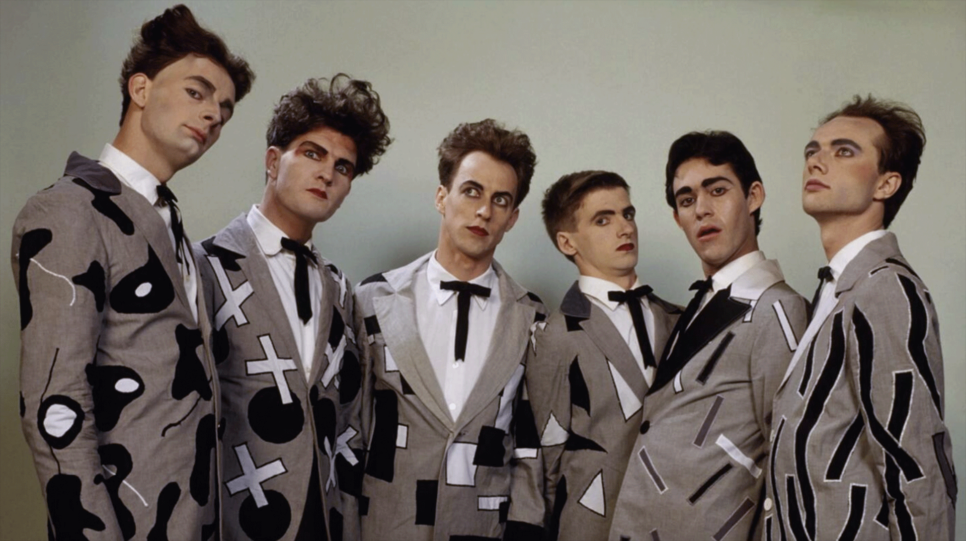 Six men wearing theatrical makeup, styled hair, and patterned grey suits with black ties pose for a portrait against a plain background.