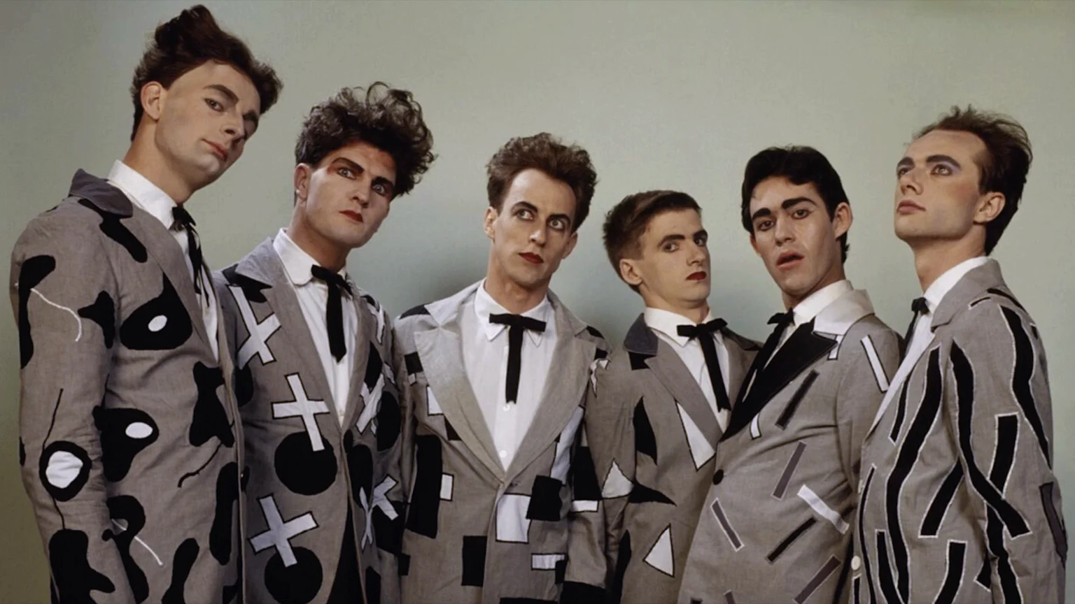 Six men wearing theatrical makeup, styled hair, and patterned grey suits with black ties pose for a portrait against a plain background.