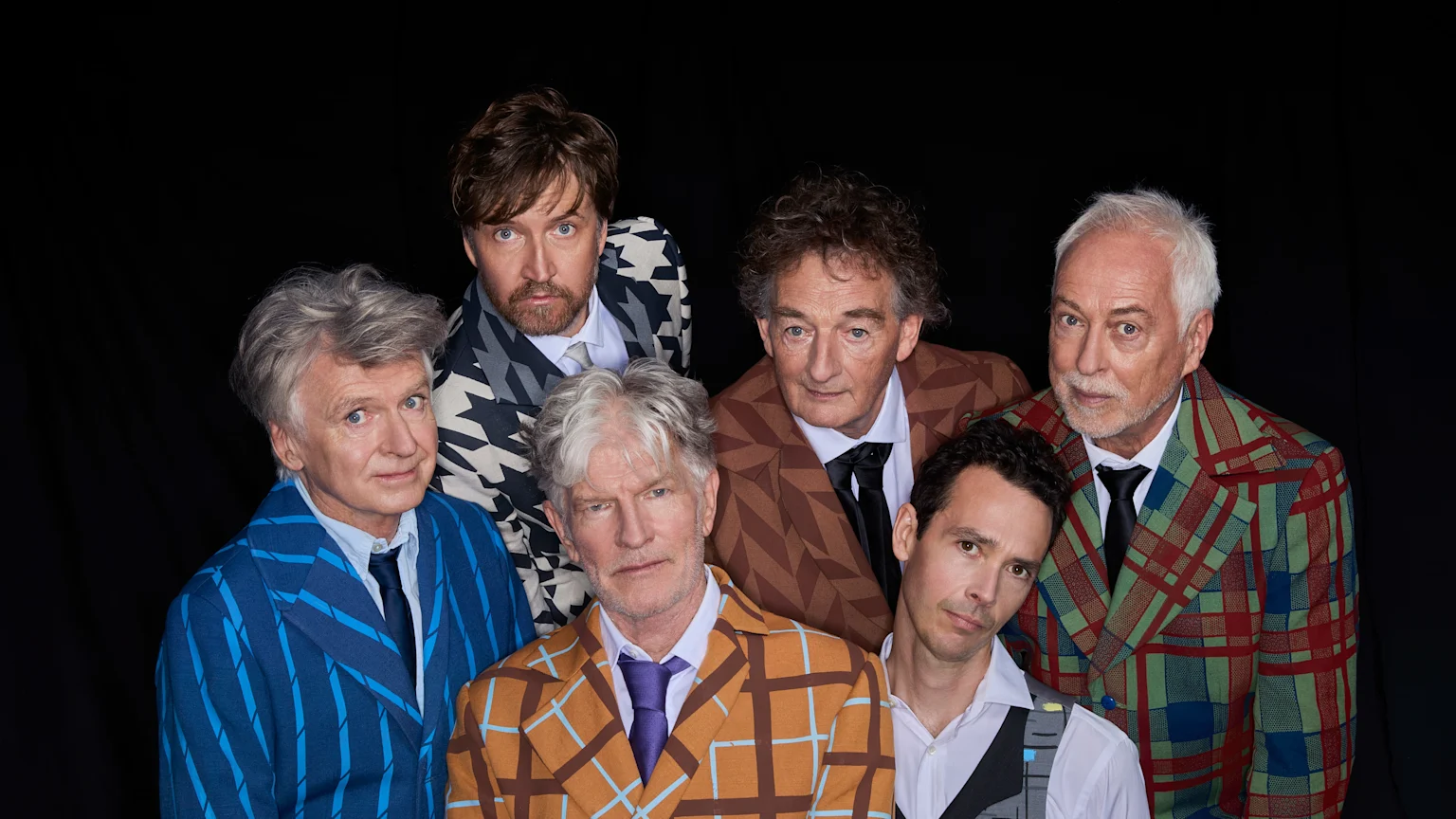 Six men wearing theatrical makeup, styled hair, and patterned grey suits with black ties pose for a portrait against a plain background.