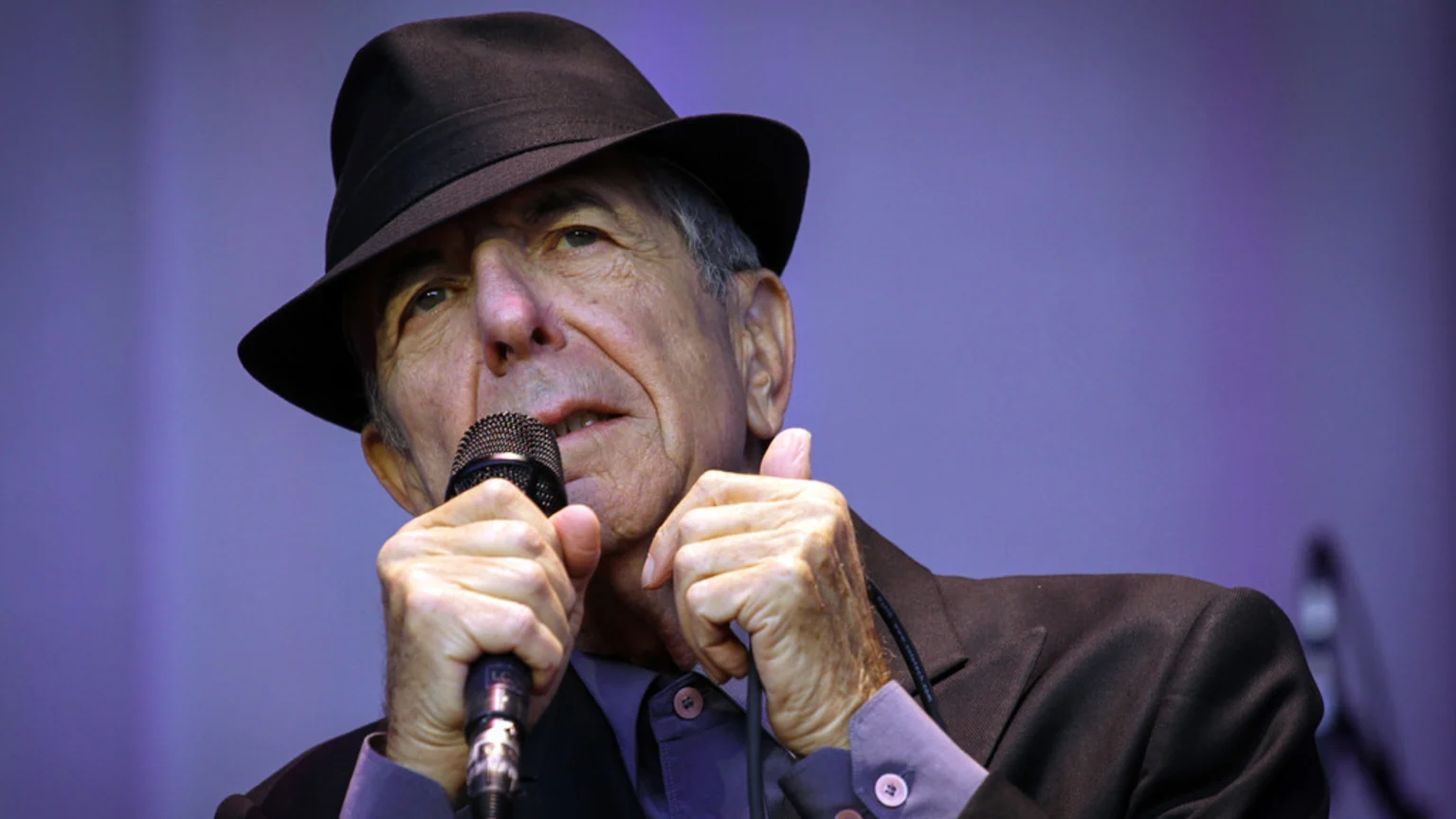 Leonard Cohen, wearing a fedora and suit, holds a microphone with both hands, appearing to sing or speak against a blurred purple background.