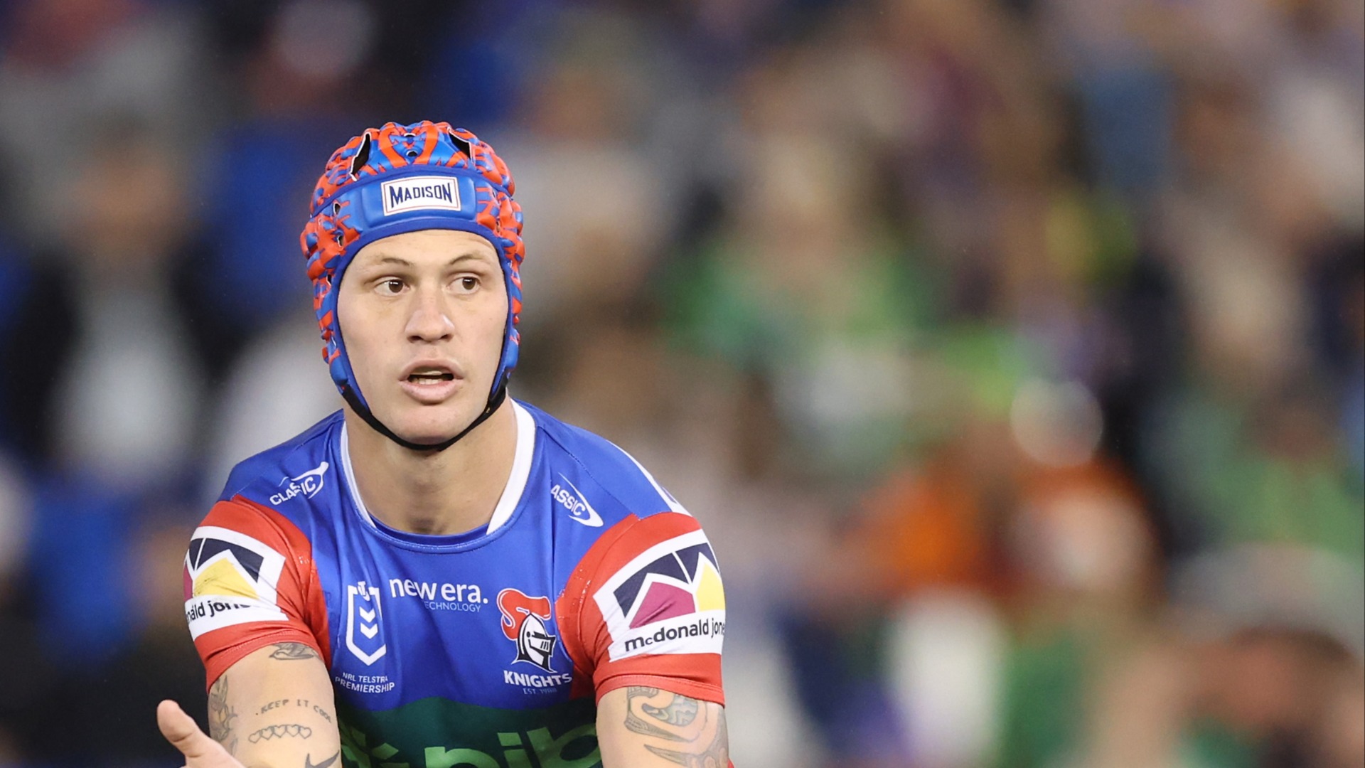 A male rugby player, Kalyn Ponga, wears a blue and red jersey and a red, blue, and orange patterned headguard with "MADISON" written on it, looking intently to his left.