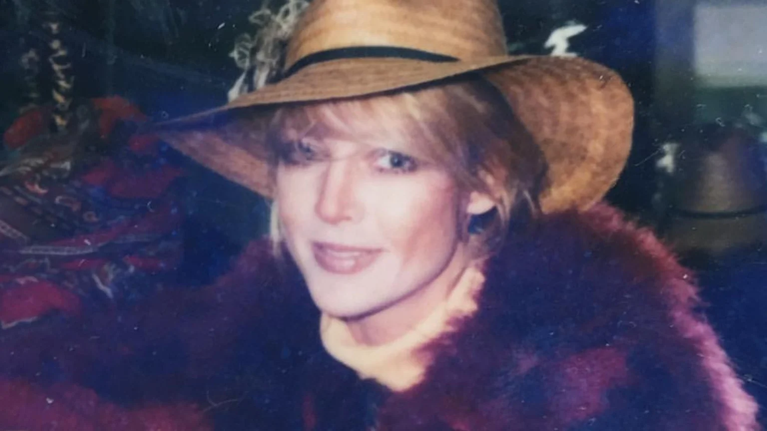 A blonde woman in a straw hat, yellow turtleneck, and reddish-brown fur coat smiles faintly.