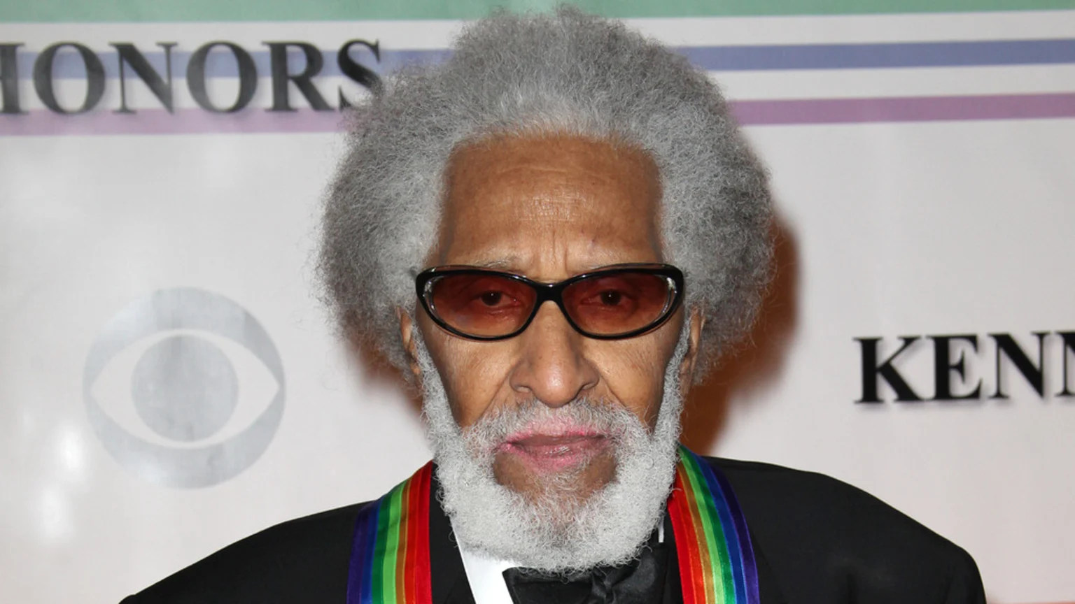 A man with a grey afro and beard wears dark sunglasses and a rainbow-striped medal, standing against a white backdrop with "HONORS", "KENNEDY", and the CBS logo visible.