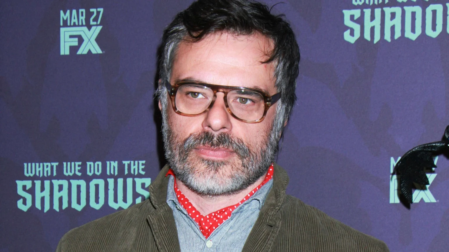 A man with a beard and glasses wears a corduroy jacket and a red polka-dot scarf in front of a purple banner for "What We Do in the Shadows" and "FX".