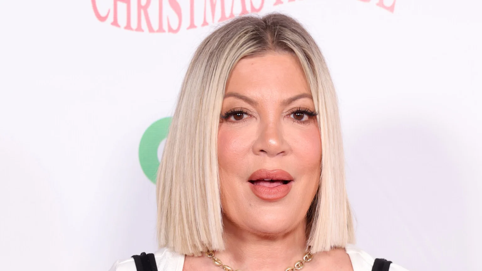 Close-up of Tori Spelling with blonde bob hair and dark roots, wearing makeup and a gold necklace, against a white background with faint red text and a green logo.