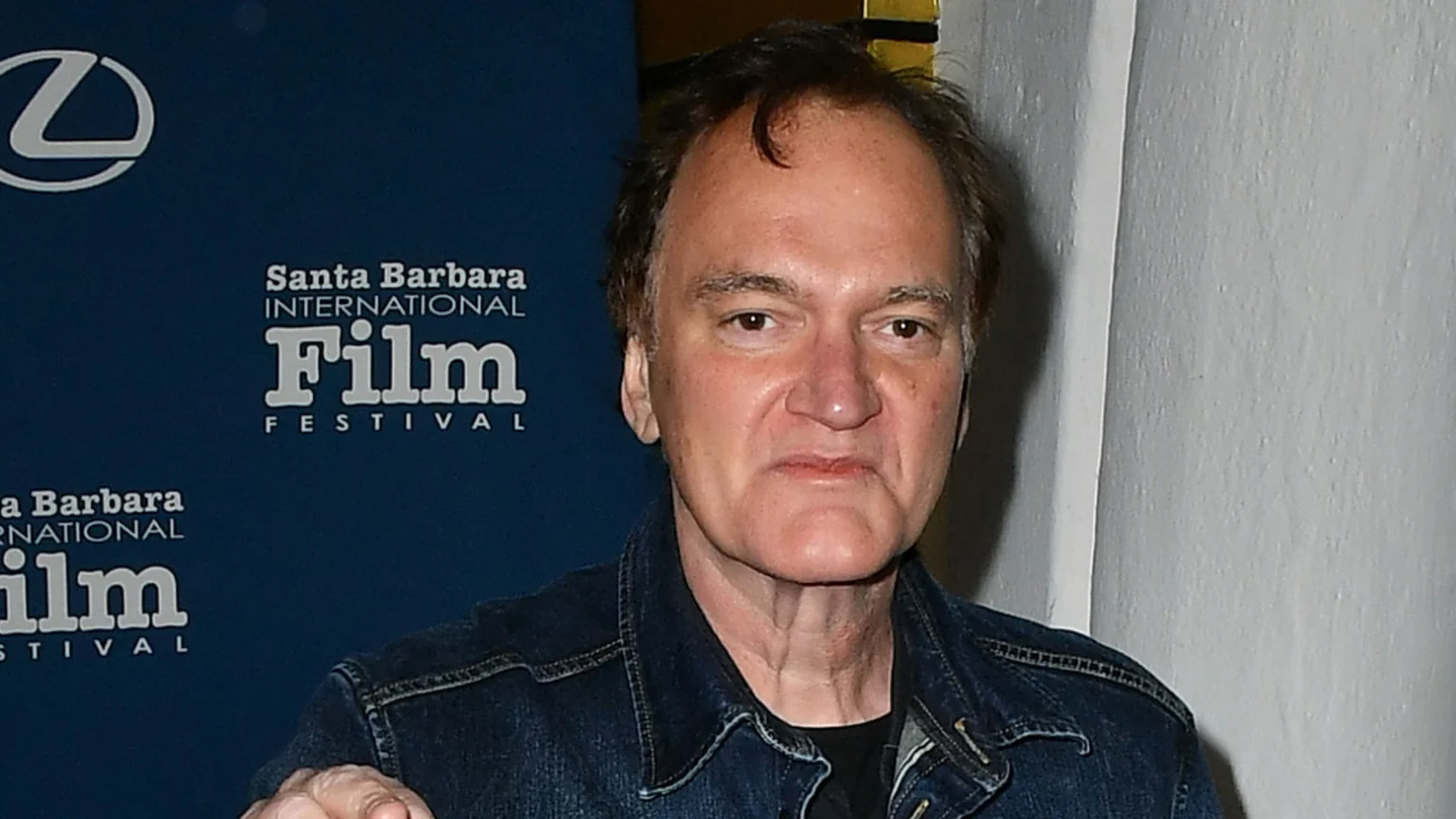 Quentin Tarantino, wearing a denim jacket, looks directly at the camera with a serious expression, with a Santa Barbara International Film Festival banner behind him.