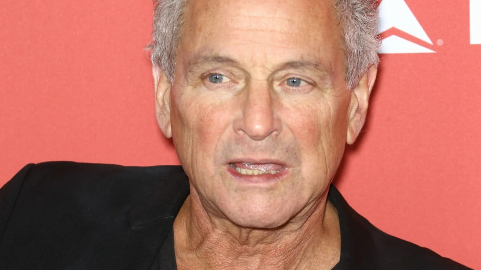 A close-up of an older man with grey hair, blue eyes, and his mouth slightly open, wearing a dark shirt against a reddish-orange background with a partial white logo.