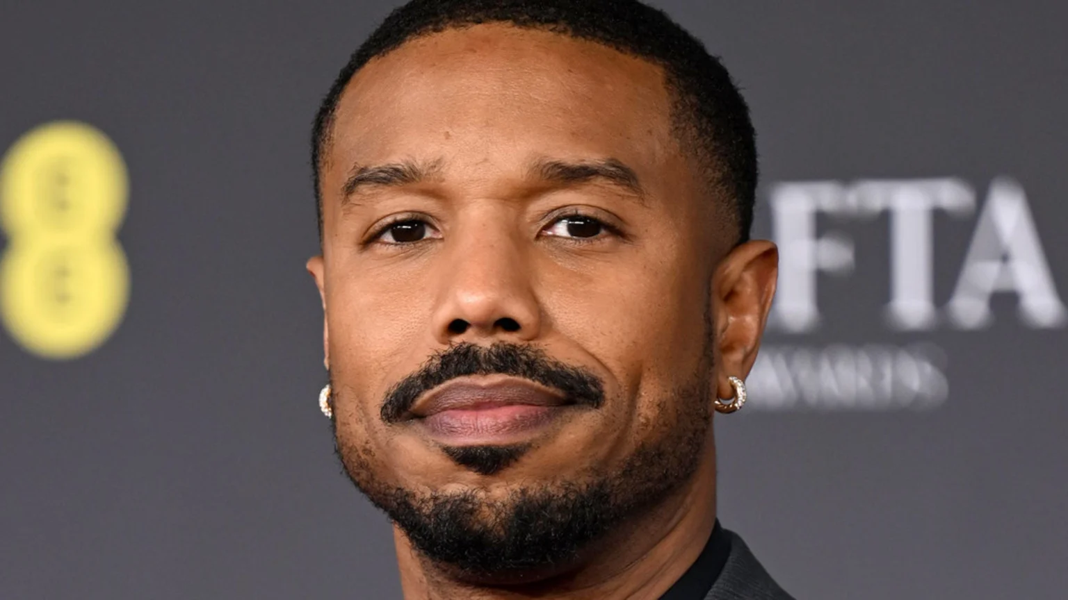 Close-up of actor Michael B. Jordan with a beard, mustache, and hoop earrings, looking forward against a dark, blurry background with faint text.
