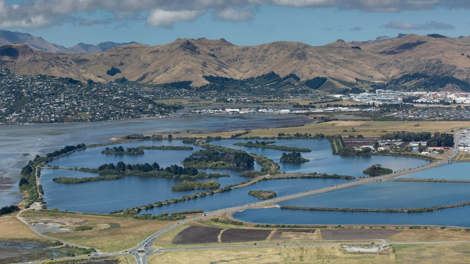 An aerial view of a landscape featuring several large, interconnected water ponds with islands and causeways, flanked by residential areas, industrial buildings, and dry hills.