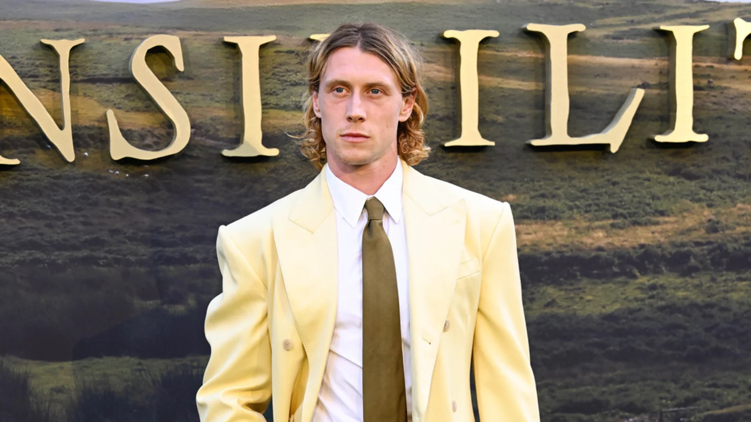 George MacKay, wearing a pale yellow suit, white shirt, and olive tie, stands in front of large golden letters against a green, hilly backdrop.